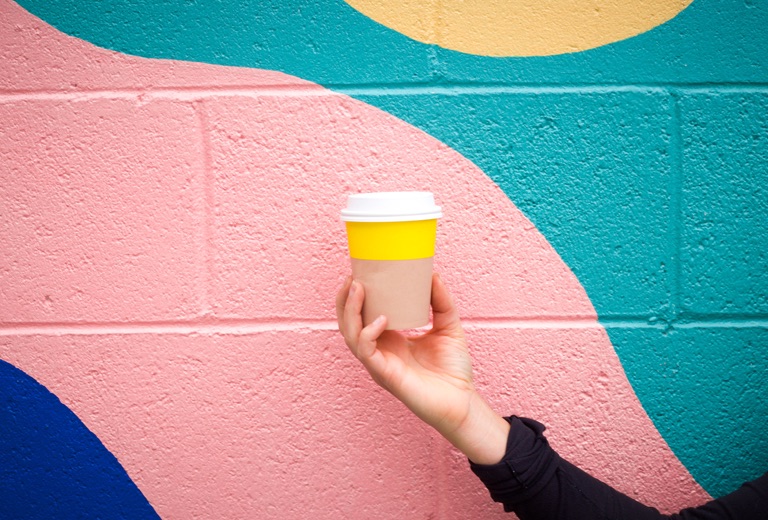 single hand holding yellow coffee cup in front of a colour block pastel graffiti stone wall