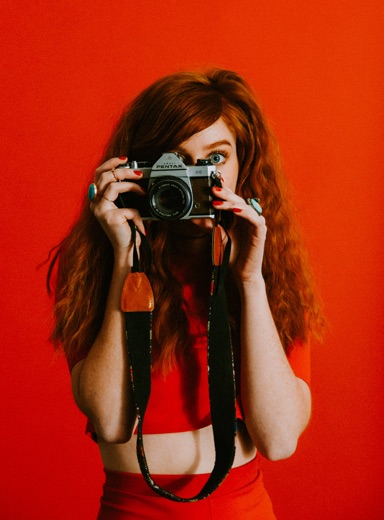 ginger-haired woman wearing red taking a photo of you with an antique camera against a red background