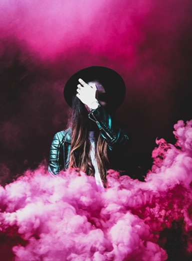 artsy woman wearing a black fedora, face covered by her hands, in a space filled with pink smoke