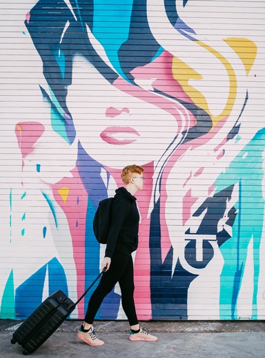 ed sheeran in all black casual style rolling a suitcase toward the right with a beautiful graffiti garage door backdrop