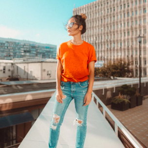 An artsy shot of Sophie wearing an orange t-shirt and blue ripped jeans with spacebuns and sunglasses coolly looking at the distance while standing on the ledge of a building