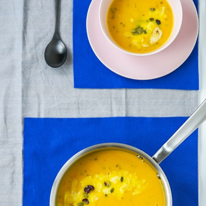 a bowl of yellow soup and a pot of the same soup on a lined table with royal blue tablecloths underneath